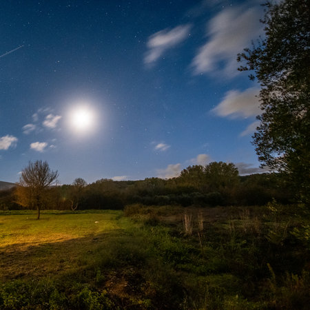 Vast night sky filled with stars and a bright full moon, with a clear satellite trail passing over a rural field and treesの写真素材