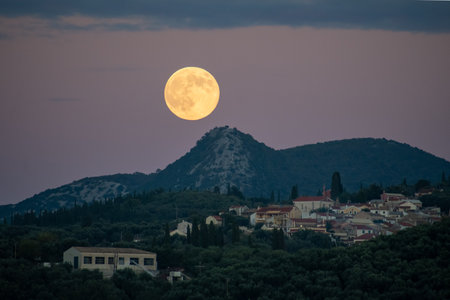 Full moon hovering directly above a mountain peak, casting a glow over a rural village and olive groves in the evening, detailed landscapeの写真素材