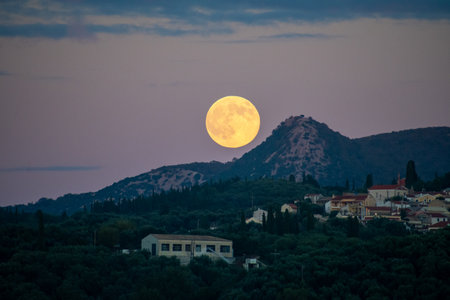 Bright orange full supermoon rising over a mountain ridge and rural Greek village with olive groves...