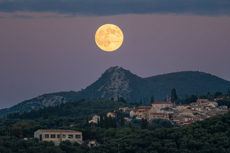 Bright yellow supermoon high in the sky over a mountain peak and a hillside village, illuminating the landscape during late twilightの写真素材