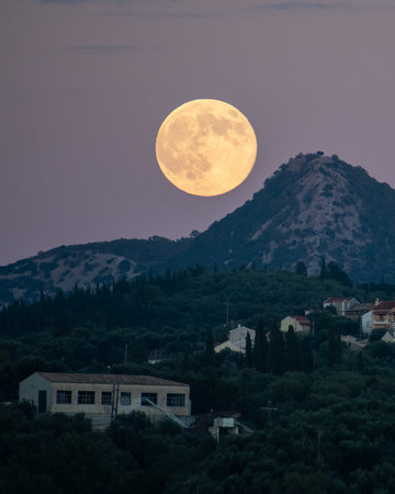 Dramatic vertical close-up of a giant full moon ascending over a mountain ridge, illuminating the silhouette of a rural Mediterranean village at dusk.の写真素材