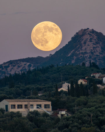 Vertical telephoto shot of a massive yellow supermoon rising behind a rugged mountain peak, looming over a small village and olive forest at night.の写真素材