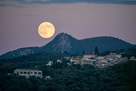 Picturesque evening landscape with a bright full moon rising next to a mountain peak, overlooking a tiered village and olive trees in the twilightの写真素材