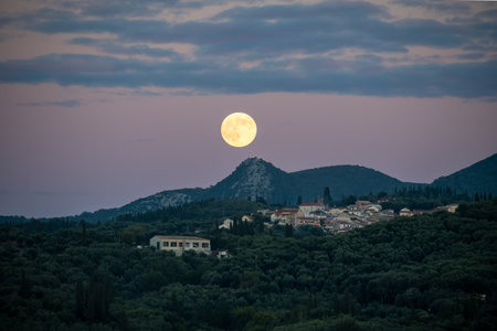 Wide landscape of a full moon glowing in a purple and pink sunset sky above a mountain peak and traditional hillside town, atmospheric evening sceneryの写真素材