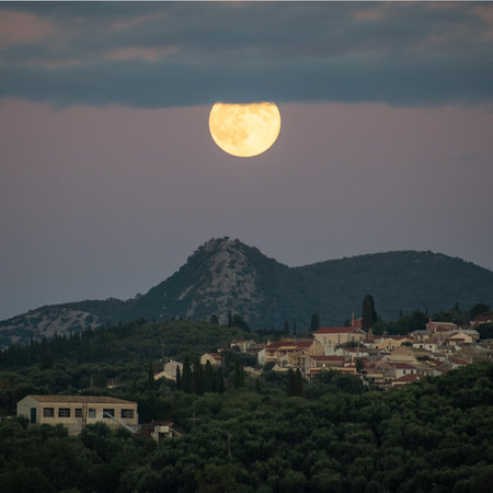 Large yellow supermoon partially obscured by a thick layer of clouds while rising over a mountain peak and rural settlement at twilight, atmospheric night sceneryの写真素材