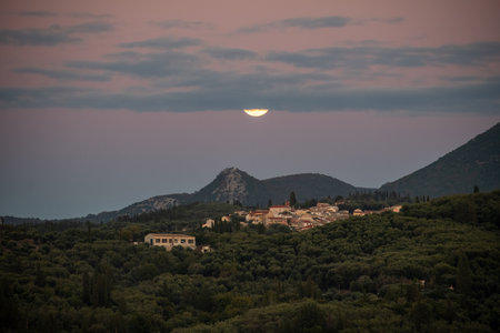 Dramatic full moon disappearing into a dark cloud bank while rising over a mountain village, creating a crescent effect in the moody evening sky over Greeceの写真素材
