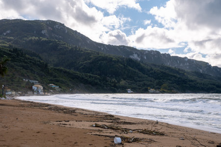 Wide angle view of a sandy beach covered in storm debris and driftwood, with rough waves and a majestic green mountain backdrop in northern Corfu, Greeceの写真素材