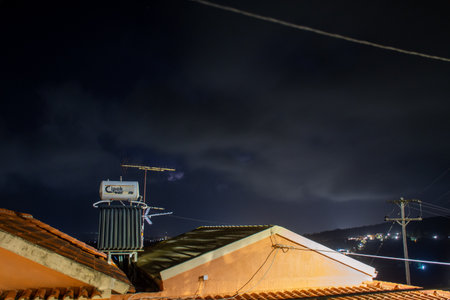 Nighttime rooftop view featuring a solar water heater and television antenna against a dark cloudy sky with distant city lights and power lines visible in the backgroundの写真素材