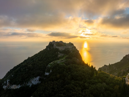 Drone Shot of Angelokastro Byzantine Fortress on Green Hilltop with Glowing Sunset and Sun Path on Sea Corfu Greeceの写真素材