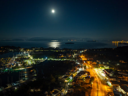 Aerial night panorama of Gouvia Marina and the Corfu coastline in Greece. A bright full moon reflects over the Ionian Sea, illuminating the harbor yachts and city street lights.の写真素材