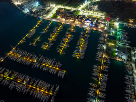 Aerial night view of a brightly lit marina harbor with moored yachts and sailboats and adjacent buildings and parking areas in Gouvia, Corfu, Greeceの写真素材