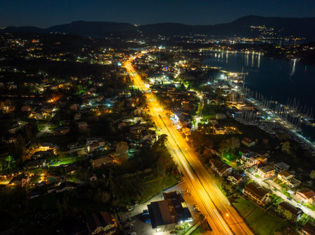 Aerial view of Corfu town outskirts at night near Gouvia, featuring illuminated streets with car light trails, residential buildings, dark coastline, and distant harbor lights in Greeceの写真素材