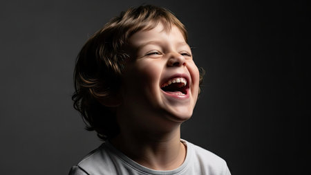 A young child is shown laughing with great enthusiasm, eyes closed, against a dark, contrasting backdrop. The image features soft lighting highlighting the child's face and expression. This portrait could be suitable for various commercial or editorial uses, conveying happiness and positive emotions.の素材