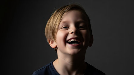 A young child is smiling broadly, their expression suggesting joy. The portrait exhibits a soft, diffused lighting with dark tones that highlight the subject. This image may be suitable for illustrating concepts related to happiness and well-being, for various commercial and editorial applications.の素材