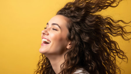 A woman with curly hair is captured mid-laugh against a yellow background. The image showcases the woman's profile, highlighting her joyful expression. The composition utilizes vibrant colors and emphasizes the hair's dynamic movement. This image could be used for various commercial or editorial projects.の素材