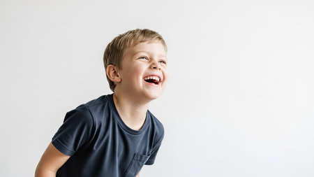 A young boy is captured mid-laugh, head tilted upward, eyes squeezed shut. He wears a dark t-shirt, set against a plain, off-white backdrop. The image conveys happiness and delight through its composition and focus. Suitable for diverse applications, including advertising and editorial content.の素材