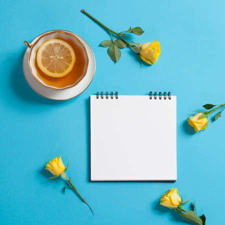 Square notepad on springs with white kraft paper, yellow rose and cup of tea are on a blue...