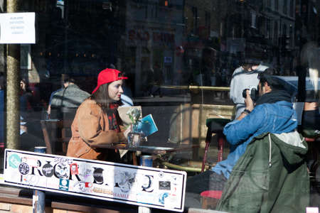 LONDON, ENGLAND - JULY 12, 2016 Man photographing his girlfriend in a red cap, sitting in a cafe....