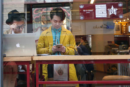 KING'S CROSS, LONDON, UK - JULY 21, 2016. Local people enjoying the cafe culture of King's Cross in London whilst drinking coffee and reading in Pret cafe. Two fashionable young man in a yellow raincoat. One of them works at a computer, other checks the pのeditorial素材
