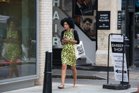 LONDON, UK - August 27, 2016: Concentrated Afro-American girl with curly phone in hand quickly goes on the sidewalk. In the background a portrait of a handsome young manのeditorial素材