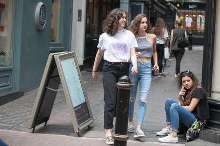 LONDON, ENGLAND - JULY 12, 2016 Two girls in jeans pass by a girl sitting on the sidewalk