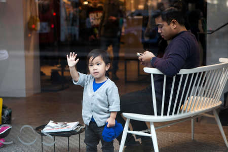 LONDON, UK - APRIL 22, 2016: The three-year Chinese child looking through a shop window on the street, while his dad sitting on the couch and waiting for wife to finish shopping.のeditorial素材