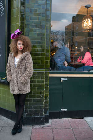LONDON, ENGLAND - JULY 12, 2016 Curly girl with long hair with an artificial butterfly in her hair. She wears faux fur coat and leggings.She leaning against wall of the houseのeditorial素材