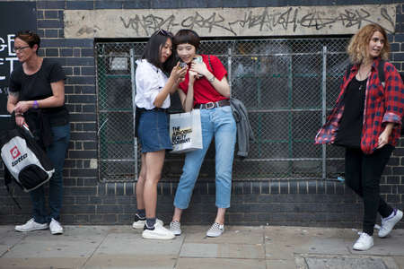 LONDON, ENGLAND - August 12, 2017 Two smiling girls are examining photos in the phone near the brick wall at Bricklaneのeditorial素材