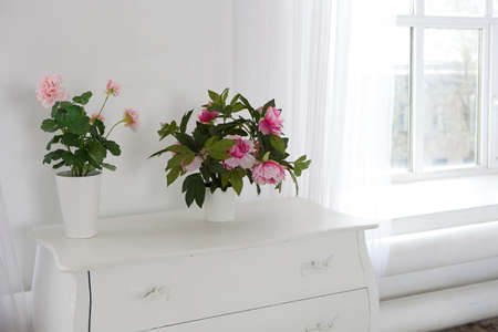 Artificial geranium and peonies in pots on a white chest of drawers as a decoration of the room