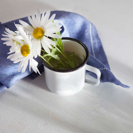 Daisies in a white enamel mug and a blue napkin on a white wooden tableの写真素材