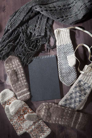 Woolen mittens, scarf and sweater with a black notepad for notes lie on the table. Copy space.