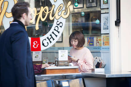 London, UK - December 22, 2017: girl in a pink sweater at a table in a cafe looks into her phone while waiting for foodのeditorial素材