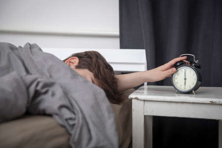 The girl, wrapped in a grey blanket, puts out her hand to turn off the alarm. There is six hours on the alarm clock.の写真素材