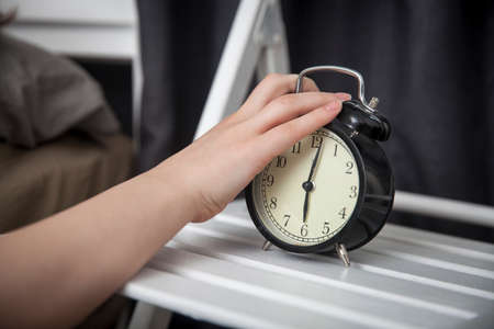 The girl, wrapped in a grey blanket, puts out her hand to turn off the alarm. There is six hours on the alarm clock.の写真素材