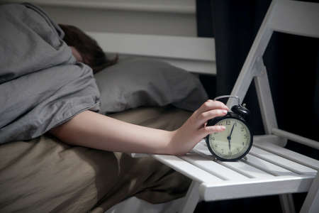The girl, wrapped in a grey blanket, puts out her hand to turn off the alarm. There is six hours on the alarm clock.の写真素材