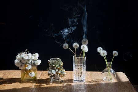 A bouquet of dandelions in a transparent glass on the wooden table opposite black backgroundの写真素材