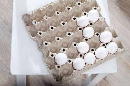 Chicken eggs in a white cardboard container lie on a beige wooden kitchen table.の写真素材