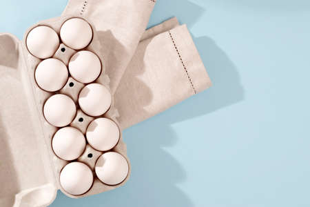 Chicken eggs in a white cardboard container lie on a blue wooden kitchen table. One egg is broken. A...