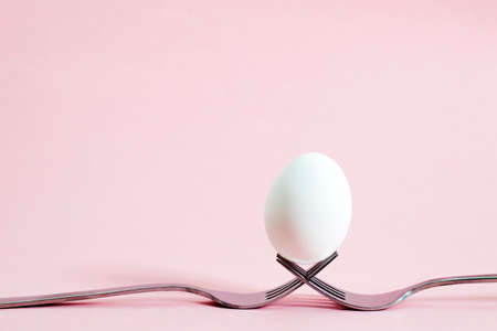 White egg lies on crossed forks on a pink background. Copy space. easter egg in balance on two...