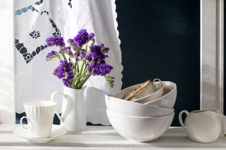 A bouquet of limonium and a set of white dishes: porcelain milk jug, coffee cup, bowls on a white rack on a dark blue background. Kitchenの写真素材