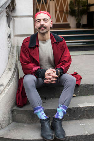 London, UK - 15 January, 2020, Street style. Hipster man with mustache in red bandana, classic burgundy cloak in the style of the seventies, gray leggings sitting on the stepsのeditorial素材