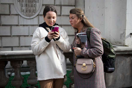 London, UK - 15 February, 2020, Fashionable people on the street . Street style. A girl in a light woolen jacket shows the news on the phone to a woman in a checked coat.のeditorial素材