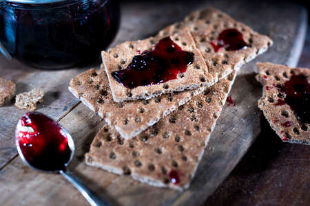 Open jar with blackcurrant jam on a wooden cutting board. Dry biscuits spread with jam, crumbs and a...