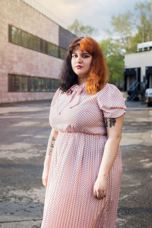 Moscow, Russia - 02 May 2020 , Street style. A young girl with red hair in a pink silk dress with a pleated skirt posing against the backdrop of an urban building.のeditorial素材