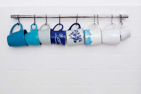 Multi-colored ceramic cups in blue tones hang on hooks above the kitchen table. Ceramic tileの写真素材