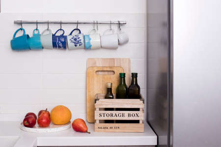 Multi-colored ceramic cups in blue tones hang on hooks above the kitchen table. Bottles with olive oil stand in a wooden box. Pears, apples and grapefruit on a round tray.の写真素材