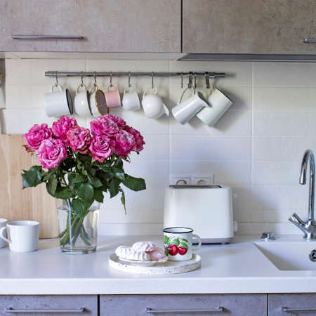 Kitchen in beige tones. Toaster. Mugs and milk jugs hang on hooks. Sink with taps. A tray with a mug and marshmallows. Bouquet of red roses in a glass vase on a white background on a wooden table.の写真素材