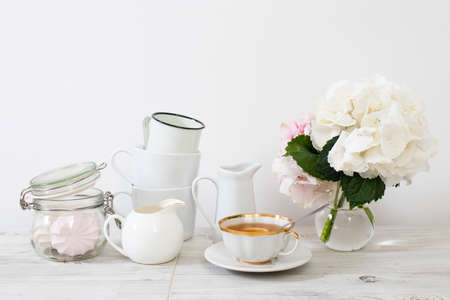 Ball-shaped white hydrangea in a round glass vase, a set of light dishes, milk jug, porcelain and enameled cups, a jug for a greeting card on a white background.の写真素材