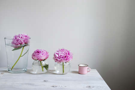 Three pink peonies in round vases on a beige wooden table against the background of a white wall. Pink cup. Space for textの写真素材