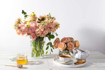 wedding bouquet of pink and orange lisianthus, antirrhinum and eucalyptus in glass vase on kitchen table. Black tea in a white cup, honey jar and spoon for honey, lemon closeup on a white backgroundの写真素材